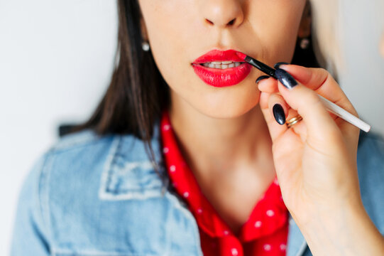 Female hand put red lipstick on lips of beautiful young brunette woman. Professional makeup
