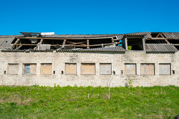 an old brick building with a ruined roof. The Windows are filled with wooden sheets. The building is set against a background of blue sky and green grass