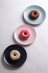 Three donuts in row on plates, chocolate, pink and vanilla donut with sprinkles, sweet glazed dessert food on white concrete textured background, angle view