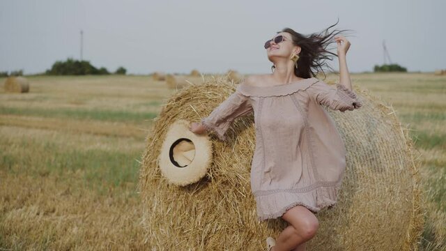 Plauful woman rejoices at haystack when holds hat and corrects hair during wind