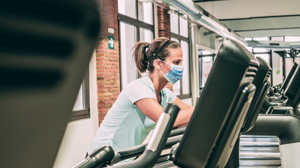 Young brunette woman does static cycling at the gym during the virus crisis. She's wearing a mask.