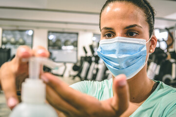 Fototapeta na wymiar Young brunette woman cleans her hands with a sanitizer gel at the gym. She's wearing a mask.