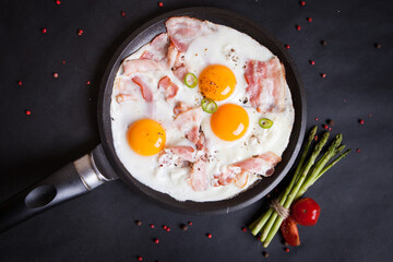 Fried eggs with three bright yolks, bacon and herbs in a black pan on a black background
