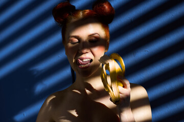 Creative portrait of a red-haired girl with a banana in her hands on a blue background with a stripe from the sun's shadow - 355212632