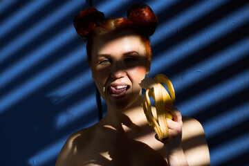 Creative portrait of a red-haired girl with a banana in her hands on a blue background with a stripe from the sun's shadow - 355212637