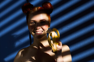 Creative portrait of a red-haired girl with a banana in her hands on a blue background with a stripe from the sun's shadow - 355212689