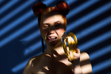 Creative portrait of a red-haired girl with a banana in her hands on a blue background with a stripe from the sun's shadow - 355212694