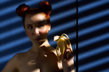 Creative portrait of a red-haired girl with a banana in her hands on a blue background with a stripe from the sun's shadow