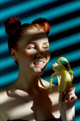 Creative portrait of a red-haired girl with a banana in her hands on a blue background with a stripe from the sun's shadow - 355213084