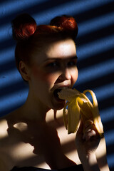 Creative portrait of a red-haired girl with a banana in her hands on a blue background with a stripe from the sun's shadow - 355213228