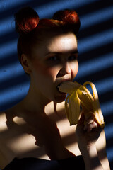 Creative portrait of a red-haired girl with a banana in her hands on a blue background with a stripe from the sun's shadow - 355213230