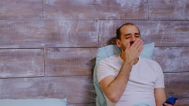 Tired man addicted to his phone, yawning and still looking at the device in his bed