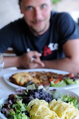 plat de nouilles, sanglier aux myrtilles et salade dans un restaurant terrasse pittoresque avec un homme content à l'arrière-plan