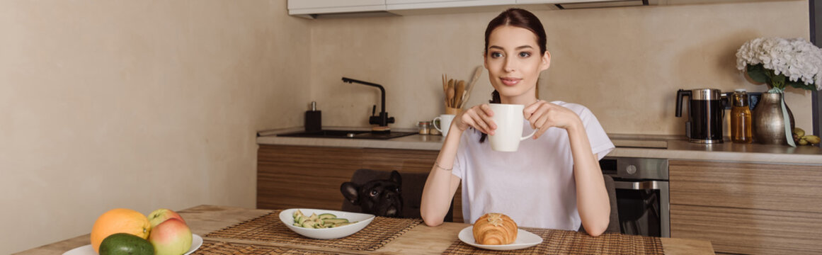 horizontal image of attractive woman holding cup of coffee near tasty breakfast and french bulldog
