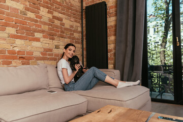 Obraz premium cheerful girl holding in arms cute french bulldog while sitting on sofa near smartphone