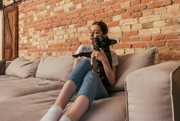 happy girl holding in arms cute french bulldog while sitting on sofa near smartphone