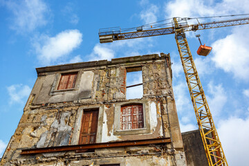 Restoration of an old building: the front wall of a house is preserved and is standing next to a yellow construction crane against white clouds in blue sky.  In the old town Miragaia Porto, Portugal. 