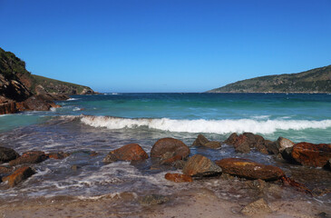 A Brazilian beach with a turquoise blue sea, a mountain with some shades of green and a clear blue sky without clouds.