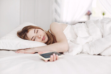 A young pretty girl in white pajamas is sleeping holding a phone in her hands.