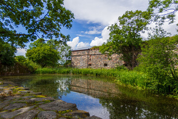 Fototapeta na wymiar The stone castle wall is embedded into the green foliage close to a pond at Suomenlinna, Finland