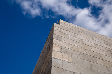 Corner view of a new large concrete brick building under construction. The stone is a tan colour with a brown mortar. The view is upward to the bright blue sky with some white fluffy clouds.   