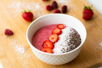 Healthy vegan trendy breakfast: magenta pink berry smoothie bowl made with frozen bananas, raspberry, strawberry and coconut milk. Topped with coconut flakes chia seeds and sliced strawberry. 