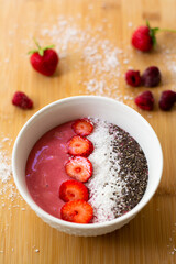 Healthy vegan trendy breakfast: magenta pink berry smoothie bowl made with frozen bananas, raspberry, strawberry and coconut milk. Topped with coconut flakes chia seeds and sliced strawberry. 