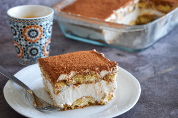 Square slice of tiramisu cake and a cup of coffee in the background, homemade gluten-free Italian layered dessert with ladyfinger biscuit and mascarpone cream decorated with cocoa powder