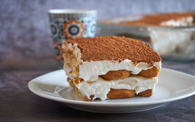 Square slice of tiramisu cake and a cup of coffee in the background, homemade gluten-free Italian layered dessert with ladyfinger biscuit and mascarpone cream decorated with cocoa powder
