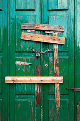 Boarded green double door, shiny and wet from rain, secured with metal chain, padlock and wooden planks. Abandoned, closed and uninhabited old house.