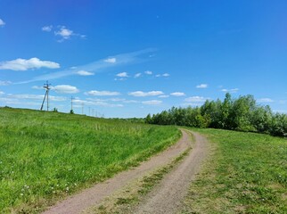 country road on a slope with green grass near a power line against a blue sky with clouds on a sunny day