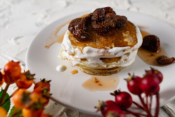 A stack of crumpets and pancakes with sour cream and strawberry sweet jam with berries on top of the dish. Dessert on a white plate with fruit in the background