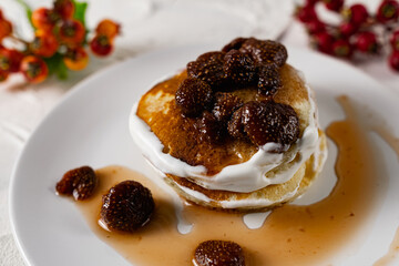A stack of crumpets and pancakes with sour cream and strawberry sweet jam with berries on top of the dish. Dessert on a white plate with fruit in the background