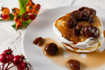 A stack of crumpets and pancakes with sour cream and strawberry sweet jam with berries on top of the dish. Dessert on a white plate with fruit in the background