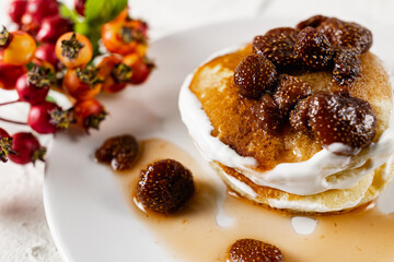 A stack of crumpets and pancakes with sour cream and strawberry sweet jam with berries on top of the dish. Dessert on a white plate with fruit in the background