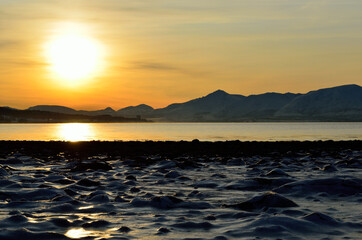 golden sunrise over blue fjord and snowy mountain with reflection on thick frozen sea shore ice