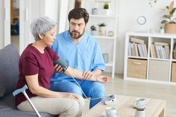 Male doctor measuring blood pressure of senior woman while they sitting on sofa at nursing house