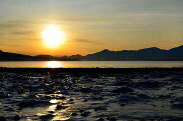 golden sunrise over blue fjord and snowy mountain with reflection on thick frozen sea shore ice