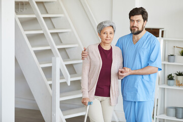 Portrait of male nurse in uniform holding for hand of senior woman and they looking at camera while standing at home