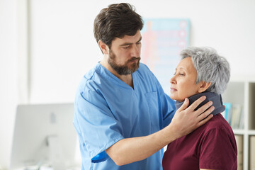Male bearded doctor examining the neck of senior woman he putting a bandage around her neck at hospital