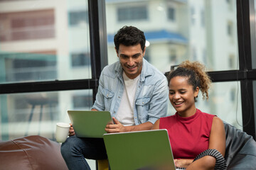 Fototapeta premium Couple of young designers sitting sofa working at modern office, two coworkers discussing fun project over a laptop and smartphone, little team of businesspeople smiling together successful meeting.
