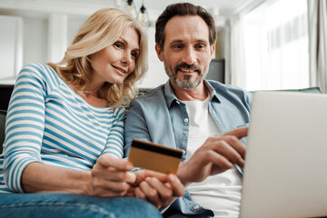 Selective focus of smiling man pointing at laptop near wife holding credit card in living room