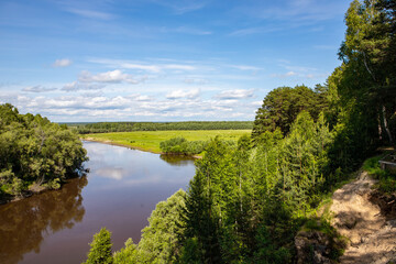Fototapeta na wymiar A small river flows around the forest, with the reflection of the trees in it against the blue sky. Nature of the Siberian expanses.