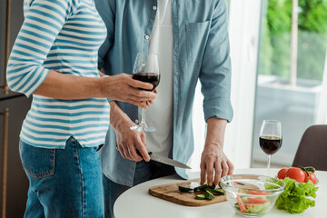 Cropped view of woman holding glass of wine near man cutting vegetables in kitchen