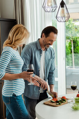 Smiling man cutting vegetables near wife with glass of wine in kitchen