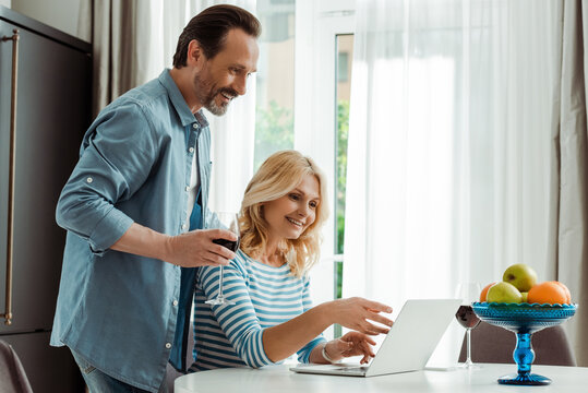 Smiling man holding glass of wine near wife pointing on laptop on kitchen table