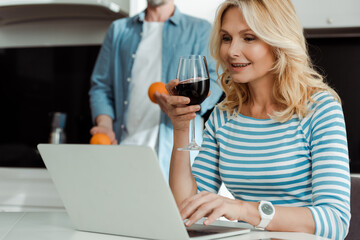 Selective focus of smiling woman holding glass of wine and using laptop near husband holding oranges in kitchen