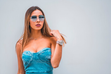 Lifestyle session, a young caucasian brunette in a blue denim dress and sunglasses on a plain gray background, with room for copy and paste, looking to the right touching her hair