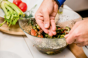 A man's hand sprinkles sesame seeds on a Greek salad. The cook adds seasoning to the vegetables
