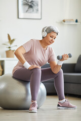 Fototapeta premium Senior woman in headphones listening to music and exercising with dumbbells on fitness ball in the living room at home